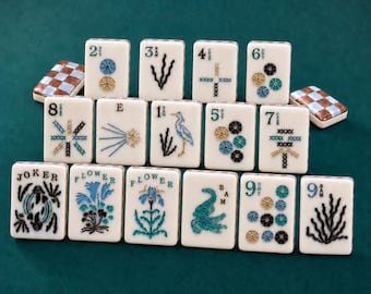 Puede incluir: Una colección de fichas de Mahjong rectangulares con caras blancas y reversos a cuadros marrones y blancos. Las fichas presentan diseños bordados en hilo azul, negro y dorado, incluyendo números, flores, un pájaro y la palabra Joker.
