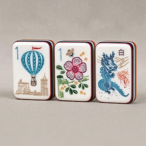 Puede incluir: Tres fichas de mahjong rectangulares con caras blancas y bordes de capas multicolores. La primera ficha presenta un globo aerost&aacute;tico azul y el n&uacute;mero 1. La segunda muestra una flor, una abeja y el n&uacute;mero 1. La tercera muestra un drag&oacute;n azul y el car&aacute;cter 白.