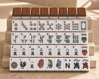 Può includere: Un set di tessere rettangolari da Mahjong disposte in file. Le tessere presentano facce bianche con illustrazioni colorate di bambù, cerchi, caratteri cinesi, fiori, un gallo e un bulldog. Il retro delle tessere superiori ha un motivo intrecciato.