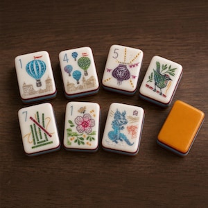 Puede incluir: Un conjunto de siete fichas de mahjong rectangulares con caras blancas e ilustraciones coloridas que incluyen globos aerost&aacute;ticos, una linterna, un p&aacute;jaro, bamb&uacute;, una flor y un drag&oacute;n, adem&aacute;s de una ficha naranja s&oacute;lida, todas con base en capas.