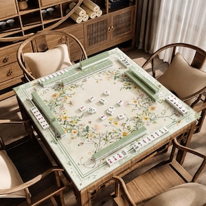May include: A square wooden mahjong table featuring a light-colored surface decorated with a floral pattern of yellow and white blossoms. The table is set with green mahjong tiles arranged in rows along the edges and scattered in the center.