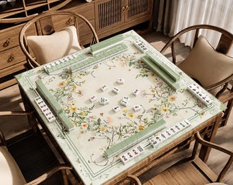 Può includere: Un tavolo da mahjong quadrato in legno con una superficie chiara decorata con un motivo floreale di fiori gialli e bianchi. Il tavolo è allestito con tessere da mahjong verdi disposte in file lungo i bordi e sparse al centro.
