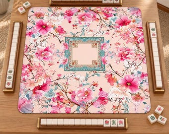 Può includere: Un tappetino da mahjong quadrato con sfondo rosa chiaro e un vivace motivo floreale di fiori rosa e bianchi. Il centro presenta un bordo quadrato decorativo in verde acqua e oro. Quattro supporti in legno sostengono le tessere bianche.