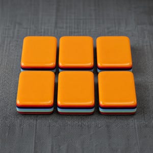 Puede incluir: Seis baldosas rectangulares dispuestas en dos filas de tres. Cada baldosa presenta una superficie superior lisa de color naranja brillante con un perfil lateral en capas que consiste en franjas horizontales en azul oscuro, azul claro, rojo y blanco.