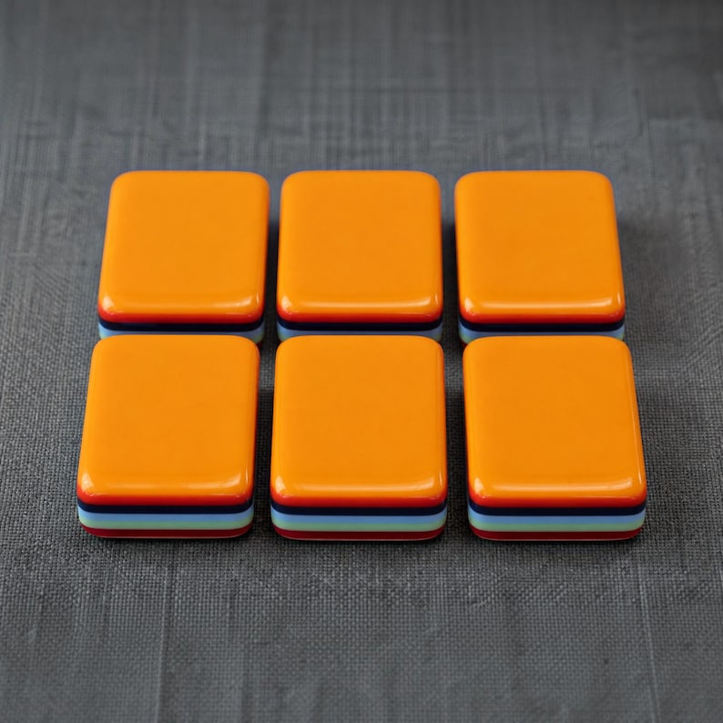 Puede incluir: Seis baldosas rectangulares dispuestas en dos filas de tres. Cada baldosa presenta una superficie superior lisa de color naranja brillante con un perfil lateral en capas que consiste en franjas horizontales en azul oscuro, azul claro, rojo y blanco.