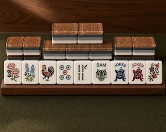 Puede incluir: Un juego de fichas de Mahjong dispuestas en un soporte de madera, con caras blancas que muestran ilustraciones coloridas de flores, un gallo, bambú, un dragón y bulldogs franceses etiquetados como JOKER. Los dorsos tienen capas de color marrón, gris y blanco.