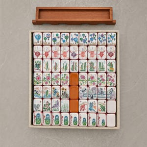 Puede incluir: Un juego de mahjong con fichas blancas que presentan ilustraciones coloridas de globos aerost&aacute;ticos, linternas, bamb&uacute;, flores y figuras, dispuestas en una bandeja rectangular con un soporte de madera separado. El juego incluye fichas numeradas.