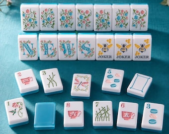 Può includere: Una collezione di tessere del Mahjong bianche con base blu, caratterizzate da motivi floreali colorati, le lettere N-E-W-S, illustrazioni di api etichettate JOKER e varie tessere numerate con icone come tazze da tè, bambù, libellule e macaron.
