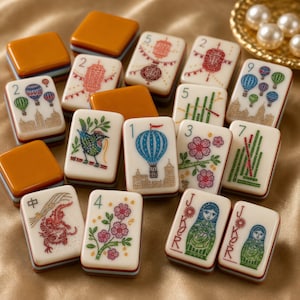 Puede incluir: Una colecci&oacute;n de fichas de mahjong rectangulares con caras de color crema e ilustraciones coloridas de globos aerost&aacute;ticos, flores, p&aacute;jaros, dragones, linternas y mu&ntilde;ecas rusas, junto a fichas con reverso naranja y marcas num&eacute;ricas.