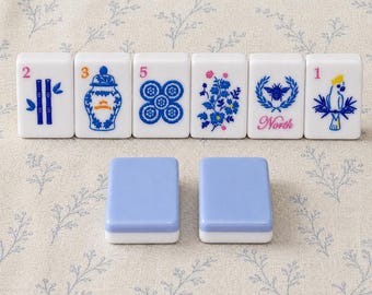 Può includere: Sei tessere rettangolari bianche da mahjong con illustrazioni blu e colorate di bambù, un vaso, motivi floreali, un'ape con la parola North e un pappagallo, accompagnate da due tessere color azzurro tinta unita con base bianca.
