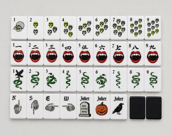Può includere: Un set di tessere bianche da mahjong con illustrazioni a tema. La riga superiore mostra teschi numerati da uno a nove. La seconda riga mostra bocche di vampiro con numerali cinesi. La terza riga mostra serpenti. La riga inferiore include mani scheletriche, una lapide, una zucca, un corvo e due tessere nere vuote.