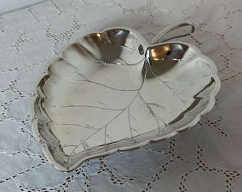 May include: A polished silver-tone metal dish shaped like a leaf. The dish features a scalloped edge and embossed vein details across the surface, with a small, curved handle extending from the top stem area of the leaf-shaped bowl.