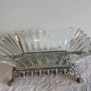 May include: A rectangular clear glass bowl with a fluted, scalloped rim design. The glass vessel is supported by a silver-toned metal base featuring ornate, decorative scrollwork and four small, curved feet that elevate the structure.