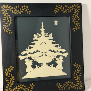 May include: A black framed wall art piece featuring a cream-colored paper cutout of a Christmas tree, two angels, and a star against a dark background. The black frame is decorated with gold-colored leaf patterns in each corner.