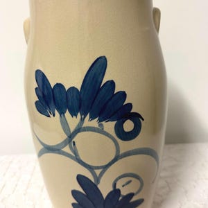 Pode incluir: Um vaso de cerâmica na cor creme com acabamento liso e vidrado e duas pequenas alças laterais. A parte frontal apresenta um design floral azul-escuro pintado à mão com pétalas estilizadas, caules em espiral e detalhes circulares no corpo.