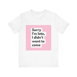 Puede incluir: Una camiseta blanca de manga corta con un gráfico cuadrado rosa centrado. Dentro del cuadrado, el texto negro dice: Sorry I'm late, I didn't want to come. La camiseta tiene cuello redondo y un corte estándar, ofreciendo un aspecto limpio y sencillo.