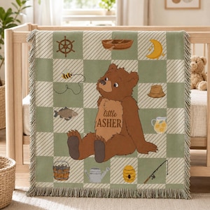 May include: A woven throw blanket with a sage green and white checkered pattern featuring a central brown bear illustration with the text little ASHER. Surrounding icons include a ship wheel, boat, moon, bee, fish, hat, pitcher, bucket, watering can, beehive, and fishing rod.