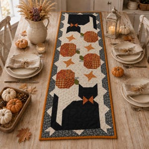 May include: A rectangular quilted table runner featuring a repeating pattern of black cats with orange bow ties, orange pumpkins, and small orange stars on a cream background, bordered by a dark gray floral print and a solid orange fabric edge.