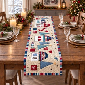 May include: A quilted table runner featuring a festive holiday design with appliqued houses, Christmas trees, hearts, and gift boxes in red, blue, and white. The runner has a cream-colored background with a multi-colored striped border.