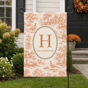May include: A rectangular garden flag featuring a cream background with an orange toile pattern of pheasants, trees, and a house. A central olive green scalloped oval contains the orange letter H and the text THE HARRISONS in orange font.