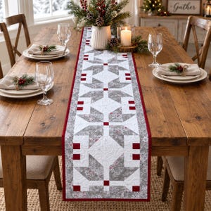 May include: A rectangular quilted table runner featuring a geometric star pattern in shades of white, gray, and dark red. The fabric displays intricate stitching, a deep red border, and small red square accents throughout the repeating design.
