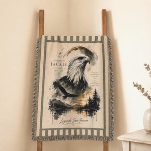 May include: A woven throw blanket featuring a central illustration of a bald eagle head and a flying eagle silhouette above a forest and lake landscape. The text reads In Memory of Jackie The Eagle 2012-2024, Thank You For Inspiring A Nation, and Legends Soar Forever.