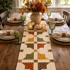 May include: A quilted table runner featuring a patchwork pattern of autumn-colored leaves in shades of orange, yellow, green, and brown on a cream background, displayed on a wooden table with white plates, green glassware, and white pumpkins.