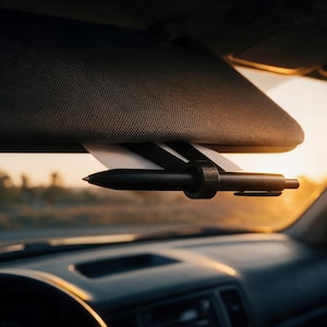 Könnte beinhalten: Ein schwarzer Stiftehalter-Clip an einer Auto-Sonnenblende, der einen schwarzen Kugelschreiber und ein weißes Blatt Papier hält. Der Clip besteht aus dunklem Material und fixiert den Stift horizontal an der Unterseite der Sonnenblende.