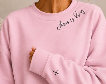 May include: A light pink crewneck sweatshirt featuring the black cursive text Jesus is King printed on the upper left chest area and a small black cross symbol printed on the left sleeve cuff near the wrist.