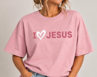 May include: A dusty rose short-sleeved t-shirt featuring the text I HEART JESUS in a bold, dark pink font. The letter I is followed by a white, hand-drawn style heart icon, and the word JESUS is printed in a clean, sans-serif typeface.