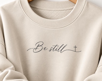 May include: A beige crewneck sweatshirt featuring the black text Be still in a cursive script font, followed by a small cross symbol. The garment has a ribbed collar and long sleeves, presented on a wooden hanger.