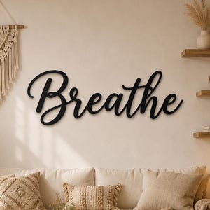 Könnte beinhalten: Ein schwarzes Wand-Schild mit dem Wort "Breathe" in einer geschwungenen Schreibschrift. Das Schild ist an einer hellen Wand über einem Sofa angebracht, zwischen einem Makramee-Wandbehang und Holzregalen mit verschiedenen Dekorationsgegenständen.