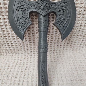 May include: A dark gray double-bitted axe featuring intricate embossed Celtic knotwork patterns on both blade faces. The handle is wrapped with textured bands and includes a decorative geometric carving along the lower shaft section.