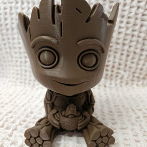 May include: A brown, 3D-printed planter shaped like a small, seated humanoid character with large eyes, a smiling expression, and a textured, tree-like head. The figure has a hollow top opening and holds a small, detailed creature in its lap.