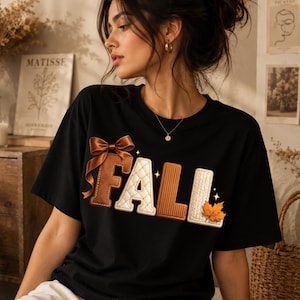 以下が含まれることがあります： 黒の半袖Tシャツで、大きく質感のあるマルチカラーの文字で「FALL」と書かれています。Fはサテンのリボンが付いた茶色、Aはキルティング加工されたクリーム色、Lはリブ編みの茶色、2つ目のLはオレンジ色のカエデの葉が付いたクリーム色です。