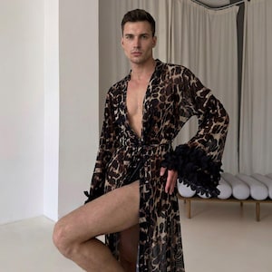 May include: A long, sheer robe featuring a brown and black leopard print pattern. The garment has a deep V-neckline, a tie waist, and wide sleeves finished with black ruffled floral-shaped fabric trim at the cuffs, creating a layered texture.