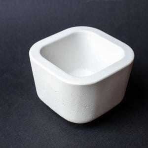 Könnte beinhalten: Ein kleiner, quadratischer weißer Behälter mit abgerundeten Ecken und einer glatten, matten Oberfläche. Das Objekt verfügt über eine tiefe, offene Mitte und einen dicken, massiven Rand mit einer porösen, strukturierten Betonoberfläche.