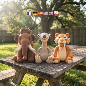 May include: Three crocheted stuffed animals sitting on a wooden surface. On the left is a brown mammoth with white tusks. In the center is a light brown sloth with large eyes. On the right is an orange and cream saber-toothed tiger with stripes.