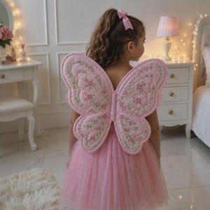May include: A pair of quilted butterfly wings in a soft pink hue, featuring intricate floral embroidery with green vines and small pink blossoms. The wings are structured with a scalloped edge and are worn over a matching pink tulle dress.