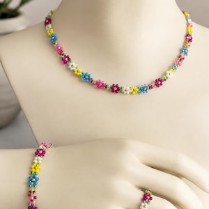 May include: A beaded jewelry set consisting of a necklace, a bracelet, and a ring. Each piece is crafted from small, colorful glass beads arranged in a repeating daisy flower pattern, featuring white, yellow, blue, and pink petals with gold centers.