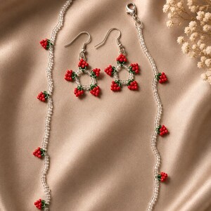 May include: A beaded jewelry set consisting of a necklace and a pair of matching earrings. The necklace features clear seed beads interspersed with small red and green strawberry-shaped bead clusters. The earrings feature star-shaped motifs.