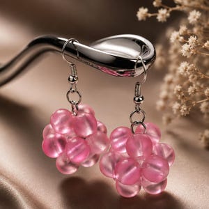 May include: A pair of dangle earrings featuring clusters of translucent pink spherical beads arranged in a rounded shape. The beads are attached to silver-toned metal fishhook ear wires with small jump rings, creating a dangling floral-like design.