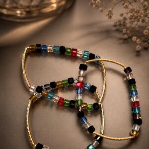 May include: Two gold-toned bracelets featuring a textured metal bar and a series of small, multi-colored cube-shaped beads. The beads are arranged in a repeating pattern of black, clear, red, green, blue, and yellow, creating a vibrant contrast.