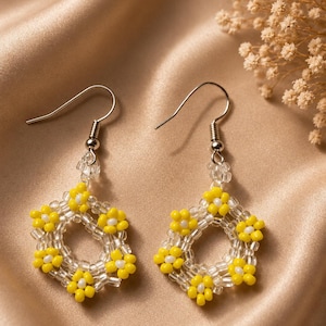 May include: A pair of dangle earrings featuring a hexagonal frame constructed from clear and yellow seed beads. Each earring showcases small flower motifs made of yellow petals and white centers, suspended from silver-toned metal fishhook wires.