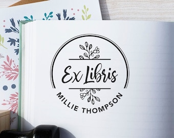 May include: A black rubber stamp with a wooden handle featuring the text Port of Reverie. The stamp impression on the open notebook page shows a circular design with the text Ex Libris and Millie Thompson, surrounded by botanical line art.
