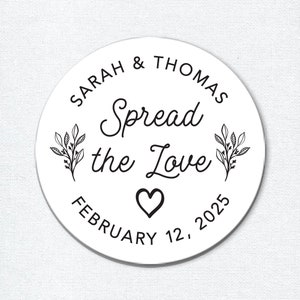 Op de afbeelding: Een witte ronde sticker met zwarte tekst en illustraties. De tekst luidt "Sarah & Thomas Spread the Love February 12, 2025". Aan weerszijden van de tekst staan twee kleine bloemillustraties.