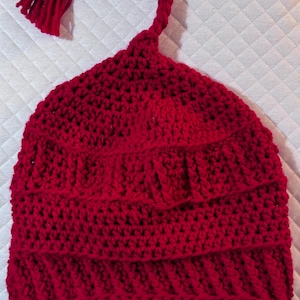 May include: A handmade red crochet hat with a unique design. The hat features a textured pattern and a decorative, braided cord that extends from the top and ends in a tassel. The hat is made of a soft, warm yarn.
