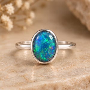 Puede incluir: Un anillo de plata con una banda de metal lisa y pulida y una gran piedra preciosa en forma de cabujón ovalado. La piedra muestra un vibrante juego de colores con destellos azul profundo y verde brillante en su superficie iridiscente.