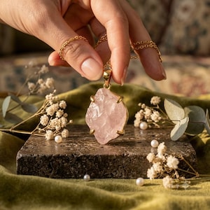 Könnte beinhalten: Eine Goldhalskette mit einem rohen, unregelmäßigen Rosenquarz-Anhänger, der von vier Krappen gehalten wird. Der Anhänger hängt an einer zarten Goldkette und zeigt die durchscheinende, blassrosa kristalline Textur des Steins in einer Goldfassung.
