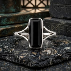 Puede incluir: Un anillo de tono plateado con una piedra negra rectangular orientada verticalmente, engastada en un bisel de metal liso. La banda presenta un diseño de brazo dividido que se estrecha al conectar con el engaste central, creando una forma simétrica.